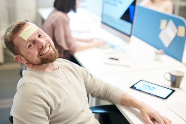 Fröhlicher Mann im Büro mit Be-Happy Notiz am Kopf